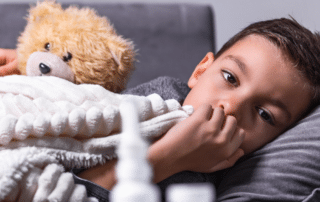 Influenza in Children | Child laying on couch, holding teddy bear and covered by a blanket. Nasal aspirator is in front of camera, out of focus.